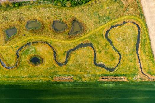 Aerial view of a meandering stream flowing through lush green meadows with small ponds and agricultural fields in Cejkovice South Moravian Region Czechia.