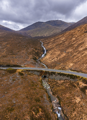 Aerial view of Eas a Bhradain waterfall cascading through a rugged mountain valley with a winding road under a dramatic cloudy sky Isle of Skye Scotland United Kingdom. Aerial view of Eas a Bhradain waterfall cascading through a rugged mountain valley with a winding road under a dramatic cloudy sky Isle of Skye Scotland United Kingdom.
