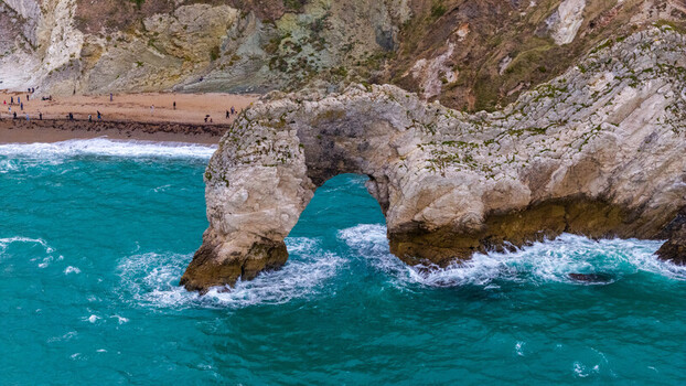 Aerial view of the natural limestone arch of Durdle Door with turquoise sea waves crashing against the cliffs and a sandy beach in the background in Wareham England United Kingdom. Aerial view of the natural limestone arch of Durdle Door with turquoise sea waves crashing against the cliffs and a sandy beach in the background in Wareham England United Kingdom.