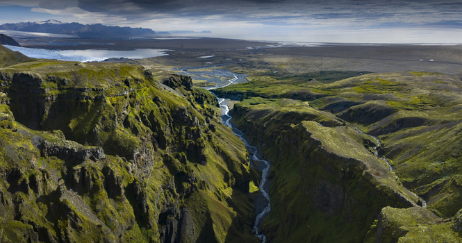 Aerial view of a lush green canyon with a winding river flowing between steep mossy cliffs towards a distant glacial lake under a cloudy sky Iceland.