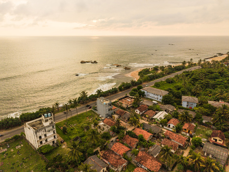 Aerial view of coastal houses with red roofs and palm trees along a seaside road by the Indian Ocean under a soft sunset sky in Galle Southern Province Sri Lanka. Aerial view of coastal houses with red roofs and palm trees along a seaside road by the Indian Ocean under a soft sunset sky in Galle Southern Province Sri Lanka.