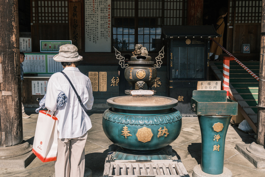 View of a woman in a hat and white shirt stands before a large ornate bronze incense burner at a temple bathed in soft light Narita Chiba Japan. View of a woman in a hat and white shirt stands before a large ornate bronze incense burner at a temple bathed in soft light Narita Chiba Japan.
