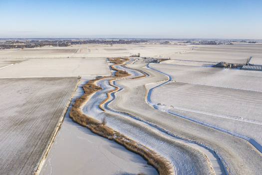 Aerial view of a winding frozen canal cutting through snow covered agricultural fields under a clear blue sky in Ursem North Holland Netherlands. Aerial view of a winding frozen canal cutting through snow covered agricultural fields under a clear blue sky in Ursem North Holland Netherlands.