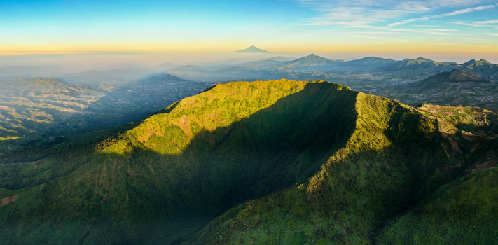 Aerial view of the lush green crater of Gunung Bismo at golden hour with distant mountain ranges under a clear sky in Kecamatan Kejajar Jawa Tengah Indonesia.