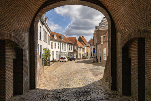 View of a historic brick archway framing a cobblestone street lined with traditional Dutch houses under a blue sky in Heusden North Brabant Netherlands.