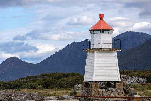 View of Laukvik fyr lighthouse with its white wooden structure and red roof standing before rugged mountains and a cloudy sky Laukvik Nordland Norway. View of Laukvik fyr lighthouse with its white wooden structure and red roof standing before rugged mountains and a cloudy sky Laukvik Nordland Norway.