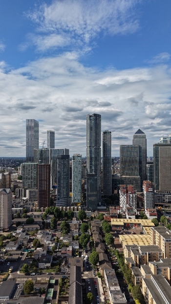 London United Kingdom 16 June 2026 Aerial view of the Canary Wharf financial district skyline with modern skyscrapers residential rooftops and a cloudy blue sky.