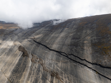 Aerial view of a steep dark grey mountain rock face with prominent geological layers and deep fissures under a cloudy sky in Nepal.