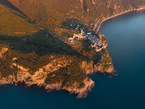 Aerial view of the village of Corniglia perched on a rocky cliff surrounded by lush green hills and the blue Mediterranean Sea at sunset in Corniglia Liguria Italy. Aerial view of the village of Corniglia perched on a rocky cliff surrounded by lush green hills and the blue Mediterranean Sea at sunset in Corniglia Liguria Italy.