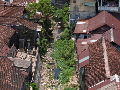 Aerial view of a narrow canal flowing between dense residential rooftops with weathered clay tiles and lush green vegetation in Kecamatan Denpasar Barat Bali Indonesia. Aerial view of a narrow canal flowing between dense residential rooftops with weathered clay tiles and lush green vegetation in Kecamatan Denpasar Barat Bali Indonesia.