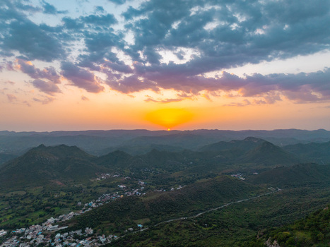 Aerial view of the sun setting over the Aravalli hills with a small town nestled in the valley below featuring rolling green landscapes and a dramatic cloudy sky in Udaipur Rajasthan India.