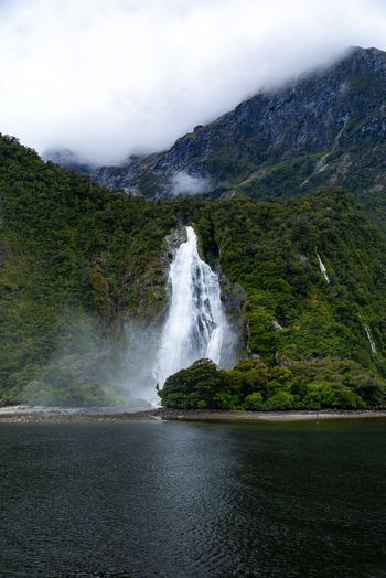 View of a powerful waterfall cascading down a lush green mountainside into the dark waters under a cloudy sky Milford Sound Southland Region New Zealand.