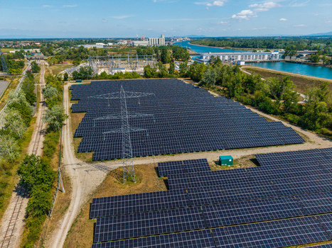 Aerial view of Solarfeld solar farm with rows of photovoltaic panels a large transmission tower and nearby industrial infrastructure along a canal in Hombourg Grand Est France.