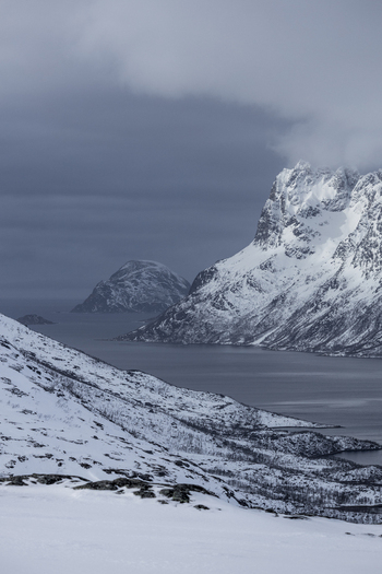 View of snow covered mountains and the cold waters of a fjord under a dark cloudy sky in winter in Ersfjord Troms Norway. View of snow covered mountains and the cold waters of a fjord under a dark cloudy sky in winter in Ersfjord Troms Norway.
