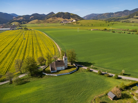 Aerial view of the Church of All Saints surrounded by vibrant yellow rapeseed and green fields with rolling mountains in the background Ludrova Zilina Region Slovakia. Aerial view of the Church of All Saints surrounded by vibrant yellow rapeseed and green fields with rolling mountains in the background Ludrova Zilina Region Slovakia.