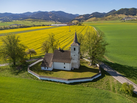 Aerial view of the historic All Saints Church surrounded by vibrant yellow rapeseed fields and green meadows with mountains in the background in Ludrova Zilina Region Slovakia. Aerial view of the historic All Saints Church surrounded by vibrant yellow rapeseed fields and green meadows with mountains in the background in Ludrova Zilina Region Slovakia.