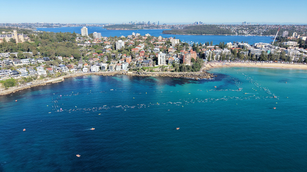 Aerial view of swimmers in the turquoise waters of Manly Beach surrounded by coastal buildings and lush greenery in Manly New South Wales Australia.