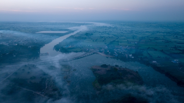 Aerial view of the Chambal River flowing through the rural landscape with a bridge crossing the water and morning fog blanketing the surrounding fields Nagda Madhya Pradesh India. Aerial view of the Chambal River flowing through the rural landscape with a bridge crossing the water and morning fog blanketing the surrounding fields Nagda Madhya Pradesh India.
