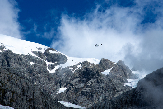 View of a small airplane flies through the expansive blue sky above rugged snow capped mountains with rocky terrain Franz Josef Glacier West Coast Region New Zealand. View of a small airplane flies through the expansive blue sky above rugged snow capped mountains with rocky terrain Franz Josef Glacier West Coast Region New Zealand.