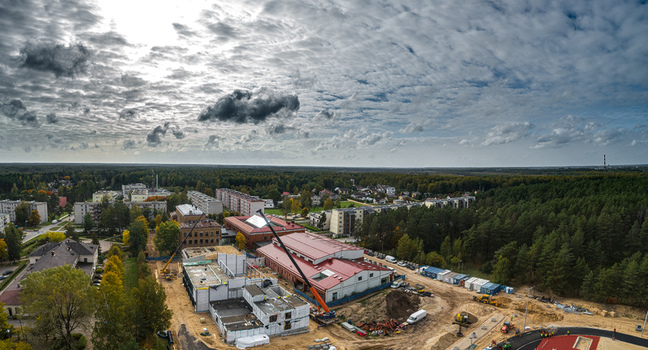 Aerial view of a construction site with cranes and building materials surrounded by residential buildings and dense forest under a cloudy sky in Latvia. Aerial view of a construction site with cranes and building materials surrounded by residential buildings and dense forest under a cloudy sky in Latvia.
