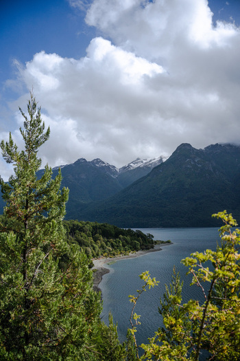 View of the serene lake and snow capped mountains within Parque Nacional Los Alerces framed by lush evergreen trees in Futaleufu Department Chubut Province Argentina. View of the serene lake and snow capped mountains within Parque Nacional Los Alerces framed by lush evergreen trees in Futaleufu Department Chubut Province Argentina.