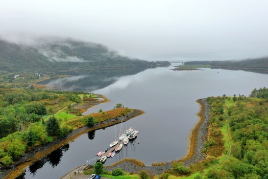 Aerial view of boats moored at a pier in a calm loch surrounded by misty mountains and lush green landscape in Glencoe Scotland United Kingdom.