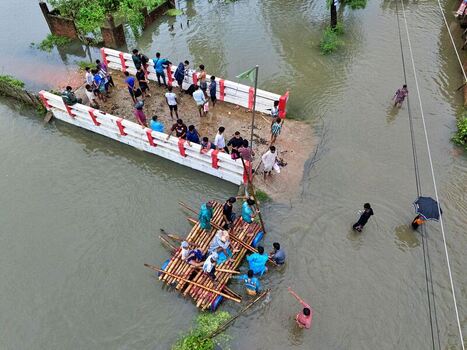Banshkhali Upazila Bangladesh 10 July 2026 Aerial view of people using a makeshift bamboo raft to navigate flooded streets near a submerged bridge with residents wading through water. Banshkhali Upazila Bangladesh 10 July 2026 Aerial view of people using a makeshift bamboo raft to navigate flooded streets near a submerged bridge with residents wading through water.