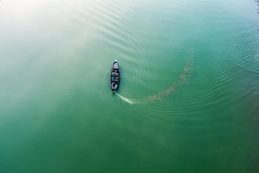Aerial view of a small boat with people moving across the calm green water of the Padma River leaving a visible wake in Rajshahi Rajshahi Division Bangladesh.