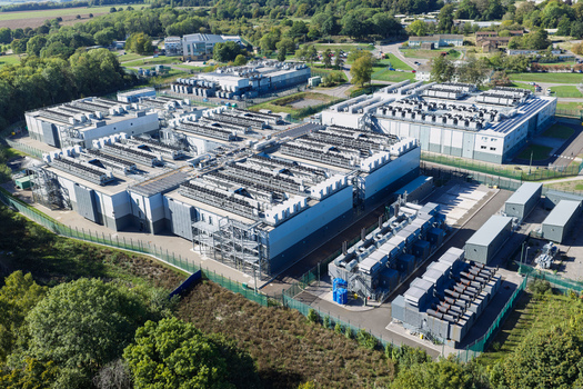 Aerial view of a large data center facility with multiple buildings rooftop cooling units and fenced perimeter in the United Kingdom.