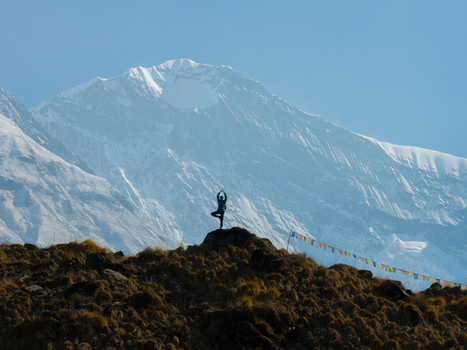 Aerial view of a person performing a yoga tree pose on a grassy hill with prayer flags against a backdrop of snow capped mountains in Nepal.