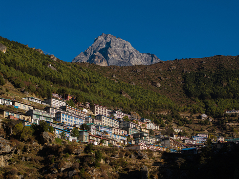 Aerial view of a mountain village nestled on a hillside with stone buildings and pine forests set against a towering snow capped peak under a clear blue sky in Nepal.