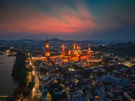 Aerial view of the illuminated Baitul Muttaqien Mosque at the East Kalimantan Islamic Center with the city skyline and Mahakam River at sunset in Samarinda East Kalimantan Indonesia.
