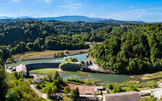 Aerial view of the Barrage de la Maigrauge dam on the Sarine river surrounded by dense green forests and rolling hills under a clear blue sky in Fribourg Fribourg Switzerland.