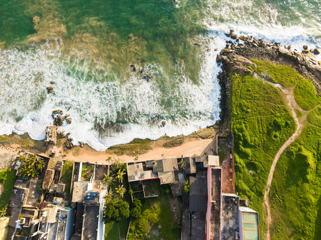 Aerial view of ocean waves crashing against a sandy beach and rocky shoreline next to coastal houses and a green grassy field Galle Southern Province Sri Lanka.