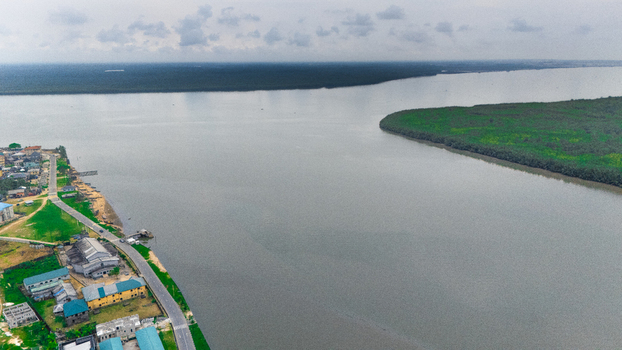 Aerial view of a river flowing past a coastal town with buildings and roads along the bank with dense green forest on the opposite side Minima Rivers Nigeria.