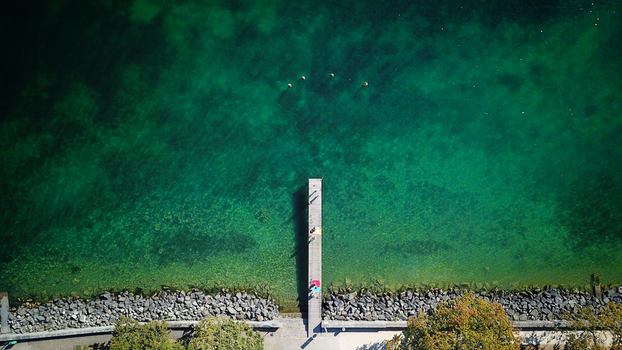 Aerial view of a wooden pier extending into the turquoise waters of Lake Geneva with people standing on the deck in Saint Prex Vaud Switzerland.