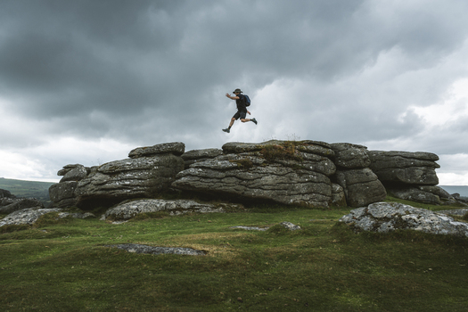 View of a man jumping on rugged granite rocks under a dramatic cloudy sky Dartmoor National Park England.