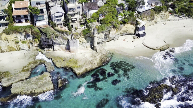 Aerial view of the historic Pirate Tower at Victoria Beach surrounded by rocky cliffs turquoise tide pools and luxury coastal homes in Laguna Beach CA United States.