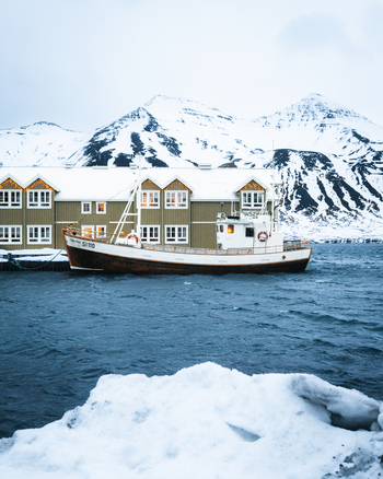 View of a fishing boat docked in the harbor of Siglufjordur with a wooden building and snow covered mountains in the background Siglufjordur Fjallabyggd Iceland.