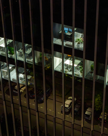 Aerial view of a modern office building in the financial center at night featuring illuminated workspaces with desks and plants and a parking area below in Warsaw Masovian Voivodeship Poland.