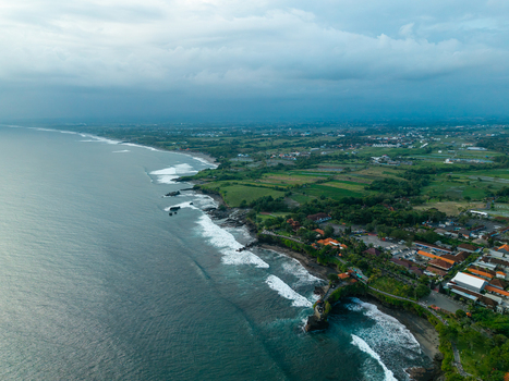 Aerial view of the rocky coastline and Tanah Lot temple area with lush green fields and ocean waves crashing against the shore in Kediri Bali Indonesia.