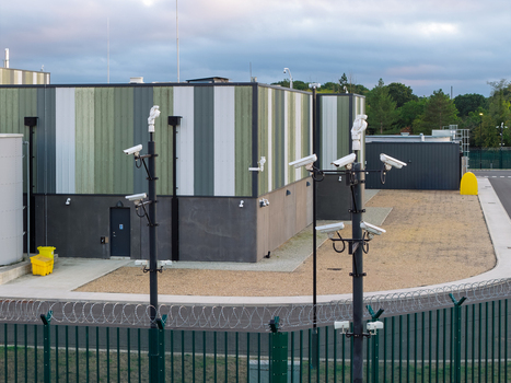 Aerial view of a data center facility featuring security camera poles perimeter fencing with barbed wire and gravel grounds in the United Kingdom. Aerial view of a data center facility featuring security camera poles perimeter fencing with barbed wire and gravel grounds in the United Kingdom.