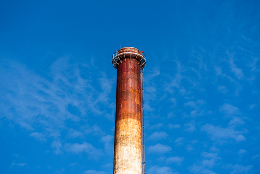 View of a tall weathered industrial chimney with rust colored textures against a bright blue sky in Poland.