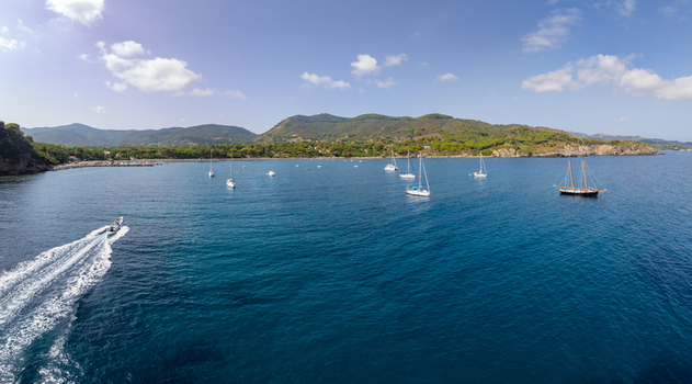 Aerial view of Lacona Beach with sailboats anchored in the blue bay and a motorboat leaving a white wake on the water surrounded by green hills in Lacona Tuscany Italy.
