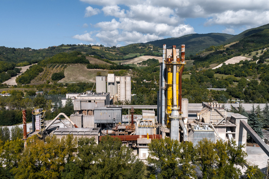 Aerial view of Sacci S.P.A. cement factory with industrial silos tall yellow kiln and metal structures surrounded by green hills and trees in Castelraimondo Marche Italy. Aerial view of Sacci S.P.A. cement factory with industrial silos tall yellow kiln and metal structures surrounded by green hills and trees in Castelraimondo Marche Italy.