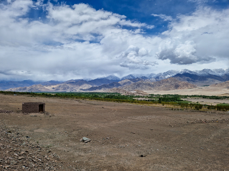 View of a small stone structure set in the arid landscape of the Himalayas with snow capped peaks under a dramatic cloudy sky in Leh Ladakh India.