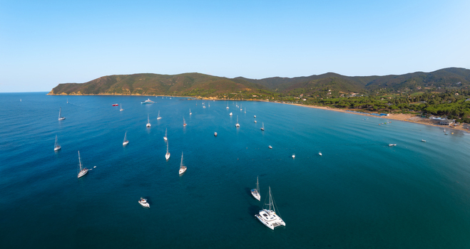 Aerial view of Lacona Beach with numerous sailboats and yachts anchored in the clear blue water surrounded by lush green hills under a clear sky in Lacona Tuscany Italy.