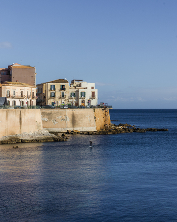 View of sun drenched old buildings with ochre walls and terracotta roofs meet the azure Ionian Sea creating a picturesque coastal scene Ortigia Sicily Italy.