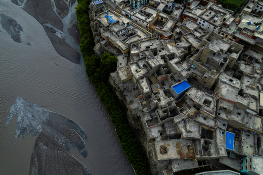 Aerial view of a traditional village with mud brick houses and flat roofs perched on a cliff edge overlooking a flowing river in Nepal.