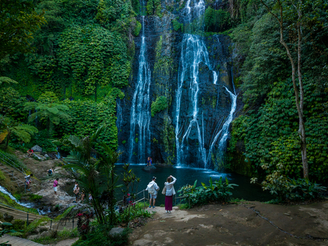Aerial view of Banyumala Waterfall cascading down a lush moss covered rock face into a clear pool with visitors standing on the bank in Sukasada Bali Indonesia.